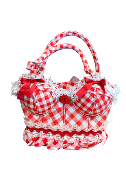 A red and white gingham handbag with ruffled trim, lace details, and a small red rose embellishment, set against a plain brown background.
