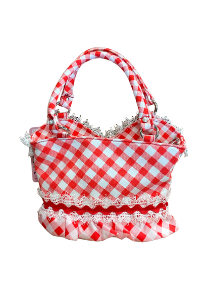 A red and white gingham checkered fabric handbag with white lace trim, featuring double top handles and a ruffled bottom edge, set against a plain brown cardboard background.