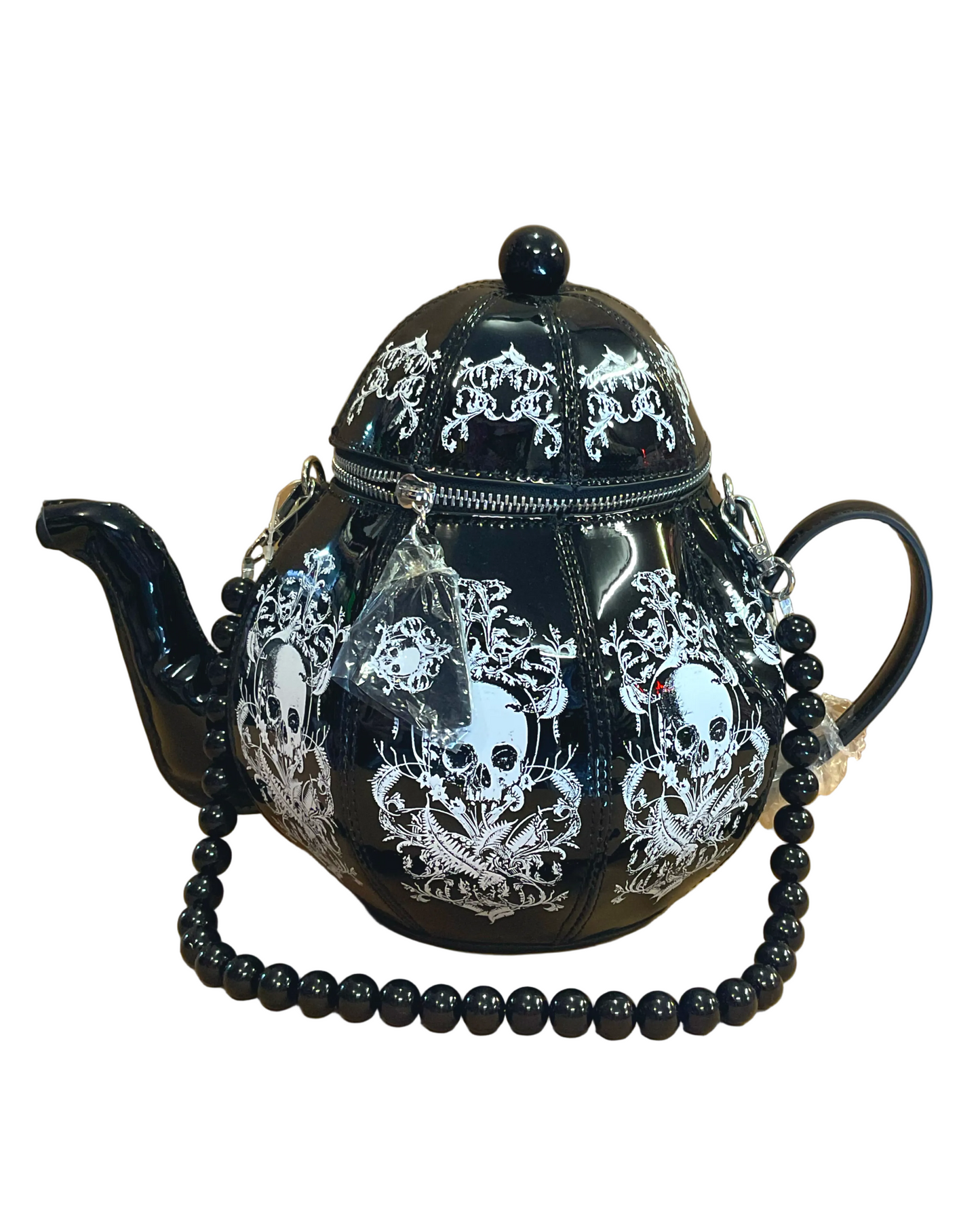 A black teapot bag with white skull and ornate gothic patterns, featuring a beaded black strap draped across the front, set against a tan background.
