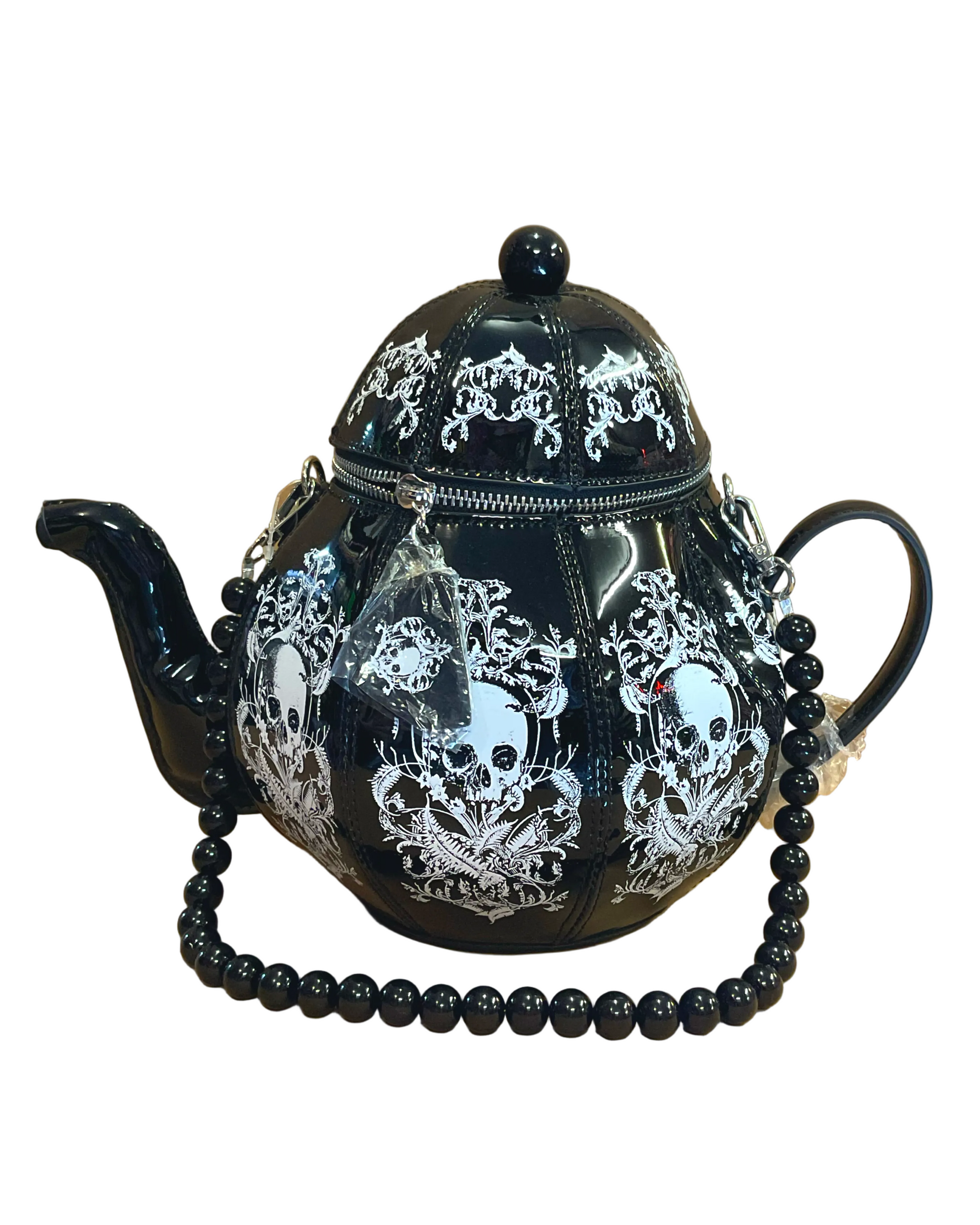 A black teapot bag with white skull and ornate gothic patterns, featuring a beaded black strap draped across the front, set against a tan background.