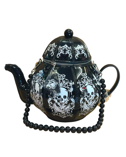 A black teapot bag with white skull and ornate gothic patterns, featuring a beaded black strap draped across the front, set against a tan background.