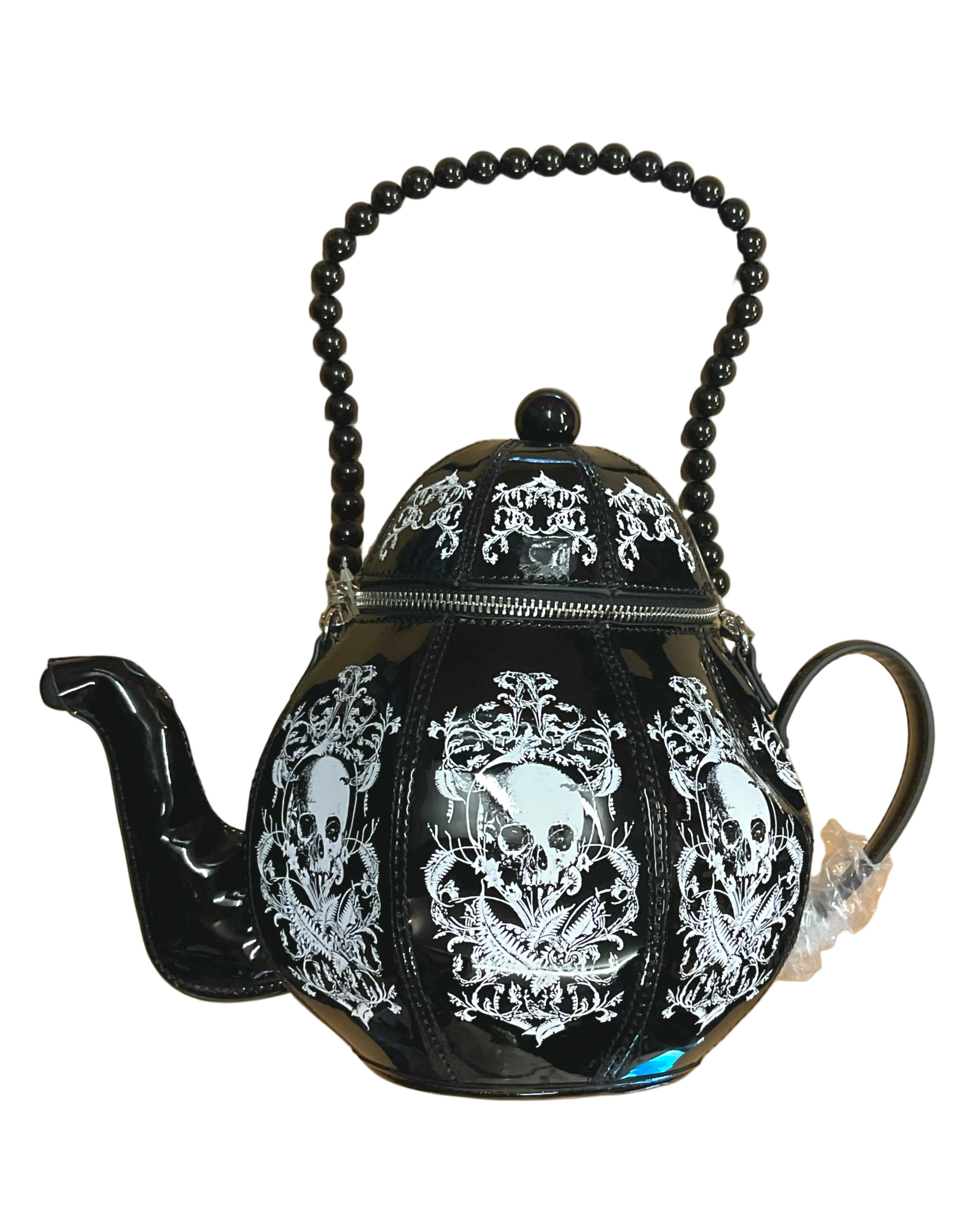 A black teapot purse with a glossy finish, decorated with white skull and baroque scroll designs. It features a beaded handle, a zipper closure, and a small white tag on the handle.